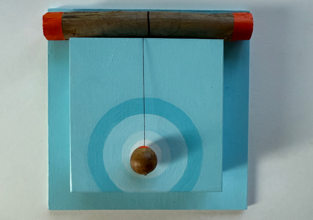 Wall-mounted assemblage of light blue painted wood, with a weathered branch capped in orange resting across the top and a small oak gall hanging by black thread over a hand-painted blue target.