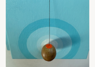 Wall-mounted assemblage of light blue painted wood, with a weathered branch capped in orange resting across the top and a small oak gall hanging by black thread over a hand-painted blue target.