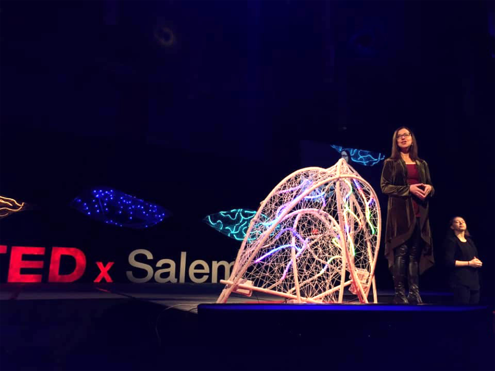 Rebecca Sira on the TEDxSalem stage beside a large PVC-and-yarn sculpture lit blue