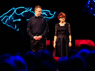 Two presenters on the TEDxSalem stage, with two of the overhead sculptures lit turquoise behind them