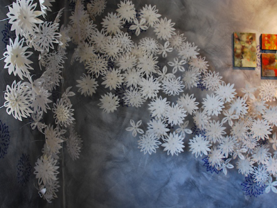Wax paper flowers cascading across a wall beside three small encaustic paintings