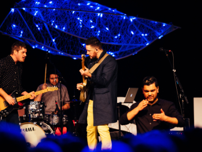 A band performing on the TEDxSalem stage, with one of the overhead sculptures lit blue above them