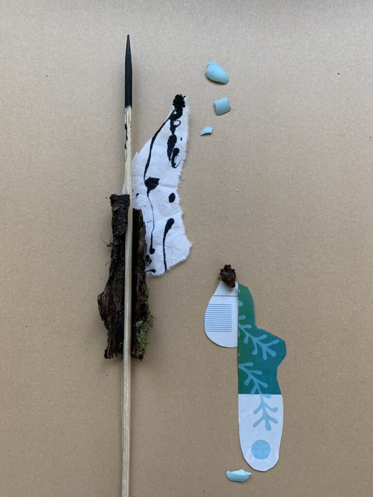 Temporary collage arrangement of a blackened wooden skewer nestled in a piece of bark, a strip of ink-marked washi paper, small turquoise robin egg fragments, cut magazine pieces, and a piece of pinecone, on cardboard.