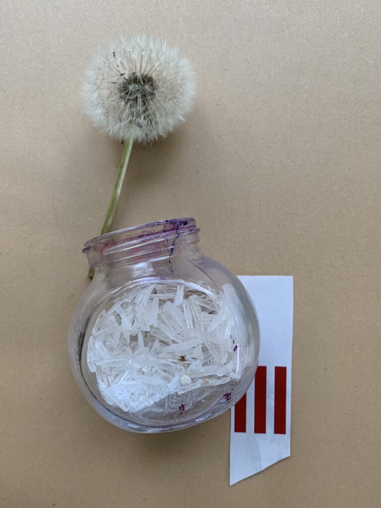 A dandelion seed head resting in the mouth of a small glass jar filled with quartz crystal fragments, beside a strip of red-and-white striped paper.