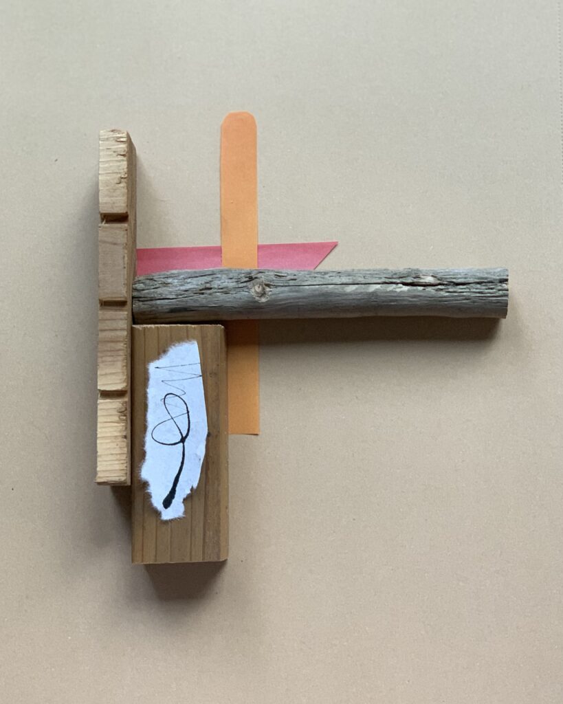 Temporary collage arrangement of a found tree branch across a wood block, a torn coral-pink paper shape, an orange paper strip, and a scrap of white washi paper marked with black ink, on cardboard.